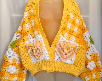 Puede incluir: Una chaqueta de punto de ganchillo amarilla con cuello en V, mangas largas y un diseño en forma de corazón en la parte delantera. La chaqueta tiene un patrón de cuadros blanco y amarillo y presenta una flor de ganchillo en cada manga.