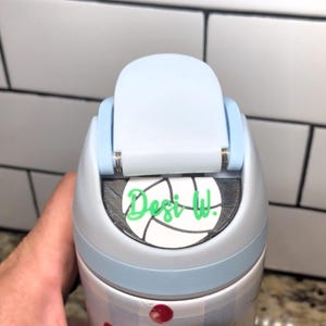 May include: A light blue and silver water bottle with a flip-top lid. The lid is light blue and has a silver hinge. The bottle has a white sticker with a volleyball design and the text "Desi W." in green.