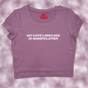 May include: A purple cropped t-shirt with the text "MY LOVE LANGUAGE IS MANIPULATION" in white. The shirt has a small red skull logo on the neck.
