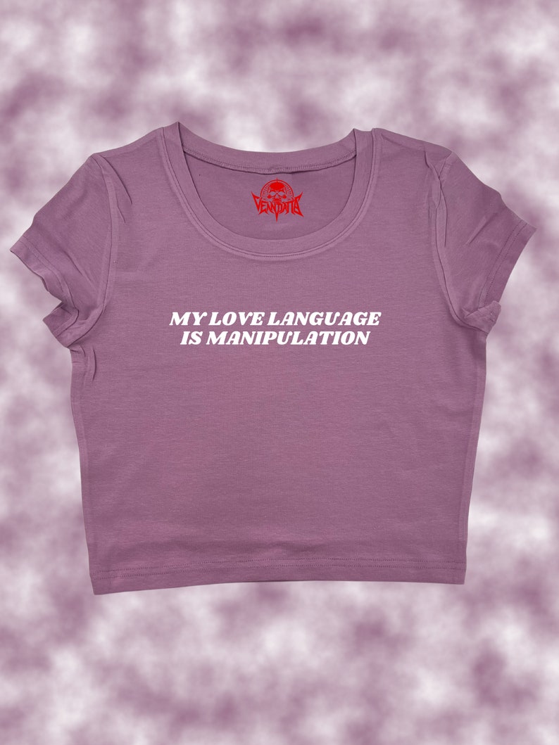 May include: A purple cropped t-shirt with the text "MY LOVE LANGUAGE IS MANIPULATION" in white. The shirt has a small red skull logo on the neck.