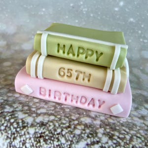 May include: A stack of three fondant books in pastel colors. The top book is green and says "HAPPY". The middle book is tan and says "65TH". The bottom book is pink and says "BIRTHDAY". White accents and diamond shapes decorate the books.