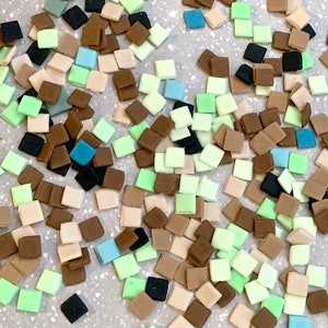 Può includere: Una collezione di piccole piastrelle quadrate di vari colori, tra cui marrone, beige, verde e blu. Le piastrelle sono sparse su una superficie grigio chiaro.