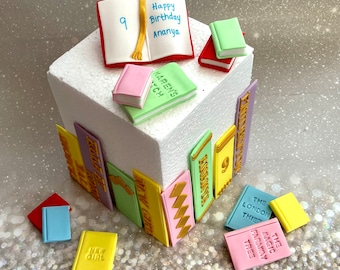 May include: A white cake decorated with colorful fondant books. The cake is decorated with the words "Happy Birthday Ananya" and the number "9".