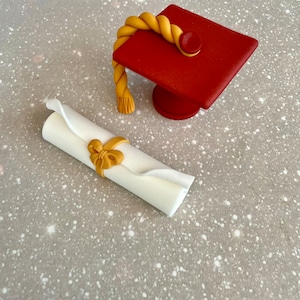 May include: A red graduation cap with a gold tassel and a white scroll tied with a gold ribbon. The cap and scroll are on a gray background.