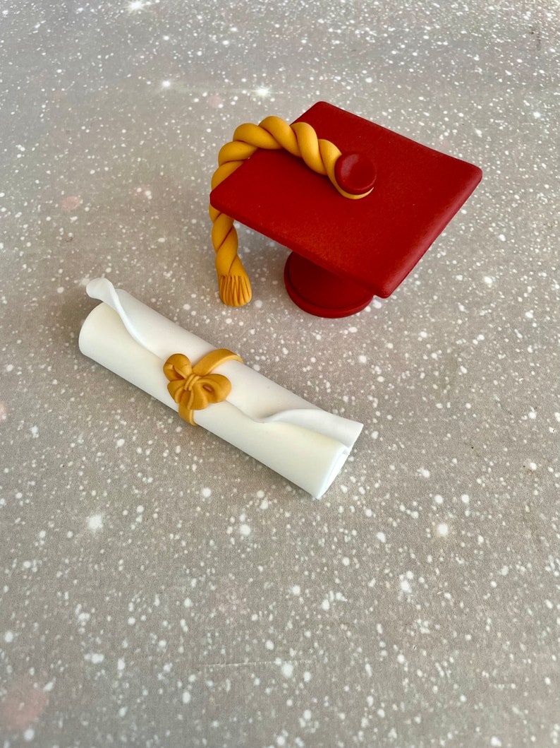 May include: A red graduation cap with a gold tassel and a white scroll tied with a gold ribbon. The cap and scroll are on a gray background.