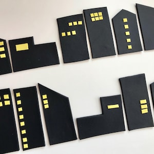 Puede incluir: Un conjunto de formas geométricas negras que representan edificios, con recortes de ventanas cuadradas amarillas. Las formas varían en tamaño y diseño, asemejándose a un paisaje urbano. Las piezas están dispuestas sobre una superficie blanca.