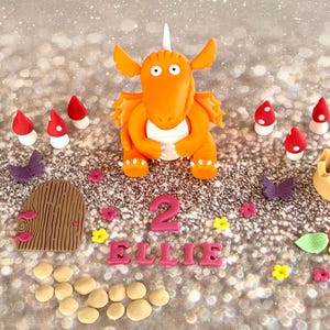 Peut inclure: Décor de gâteau en forme de dragon orange en pâte à sucre avec une corne blanche, posé sur un gâteau décoré de champignons, de fleurs, de feuilles et de pierres en pâte à sucre. Le gâteau comporte également une porte en bois, une couronne, des papillons, ainsi que le nom "Ellie" et le chiffre "2" en pâte à sucre rose.