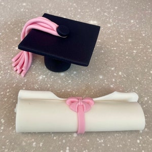May include: A black graduation cap with a pink tassel and a white diploma with a pink bow. The cap and diploma are made of fondant and are perfect for a graduation cake.