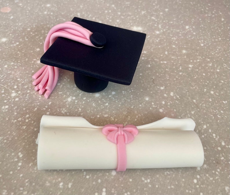 May include: A black graduation cap with a pink tassel and a white diploma with a pink bow. The cap and diploma are made of fondant and are perfect for a graduation cake.