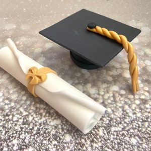 May include: A white rolled up diploma with a gold bow tied around it and a black graduation cap with a gold tassel.