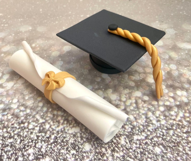 May include: A white rolled up diploma with a gold bow tied around it and a black graduation cap with a gold tassel.