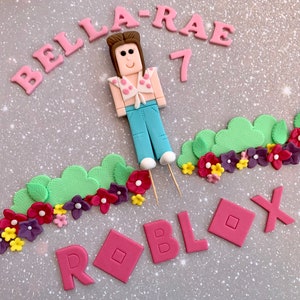 以下が含まれることがあります： ピンクと白のケーキトッパーは、ピンクのシャツと青いジーンズを着たRobloxのキャラクターを特徴としています。ケーキトッパーは、ピンク、グリーン、パープルのフォンダンフラワーと葉で囲まれています。ピンクのフォンダンには、「Bella-Rae 7」と「ROBLOX」の文字も含まれています。