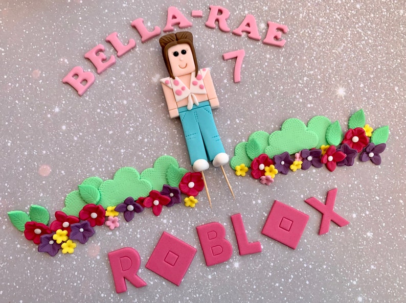 以下が含まれることがあります： ピンクと白のケーキトッパーは、ピンクのシャツと青いジーンズを着たRobloxのキャラクターを特徴としています。ケーキトッパーは、ピンク、グリーン、パープルのフォンダンフラワーと葉で囲まれています。ピンクのフォンダンには、「Bella-Rae 7」と「ROBLOX」の文字も含まれています。