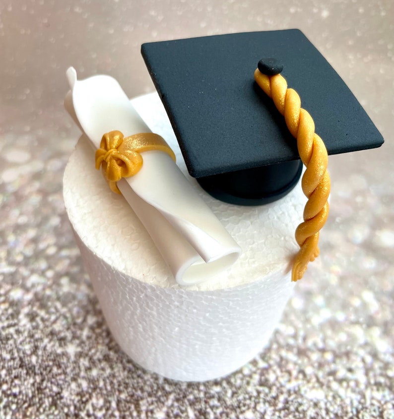 May include: A black graduation cap with a gold tassel and a white scroll tied with a gold ribbon. The cap and scroll are sitting on a white cake stand.
