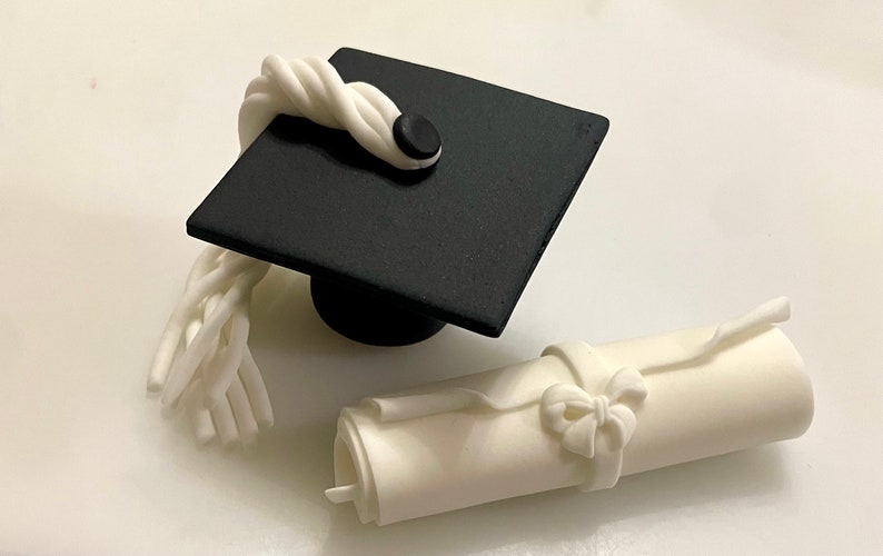 May include: A black graduation cap with a white tassel and a white scroll with a bow. The cap and scroll are made of fondant and are perfect for decorating a graduation cake.