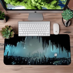 May include: A black and blue mousepad featuring a scene from the video game Hollow Knight. The design shows a character in a dark cave setting with stalactites and glowing blue light. A white keyboard and mouse are on a wooden desk.