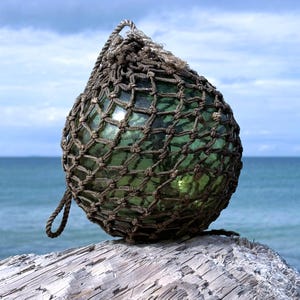 May include: A green glass fishing float encased in a brown rope net. The float is teardrop-shaped and sits on a piece of weathered wood, with the ocean and sky in the background. The net has a loop for hanging.