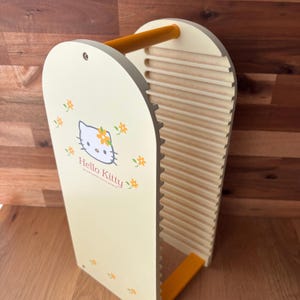 May include: A light yellow wooden organizer with a Hello Kitty graphic and floral accents. The organizer has a handle and a slotted design. The Hello Kitty graphic is centered on the front panel, with the text "Hello Kitty" below it.