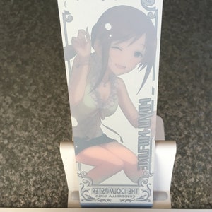 May include: A white bookmark featuring a colourful illustration of a girl with brown hair and a green and white outfit. The text "CINDERELLA GIRLS THE IDOLM@STER" is printed on the bookmark.