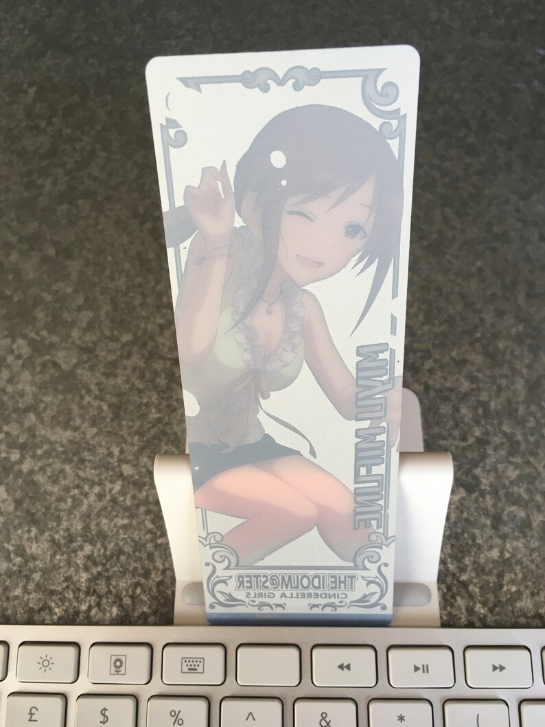 May include: A white bookmark featuring a colourful illustration of a girl with brown hair and a green and white outfit. The text "CINDERELLA GIRLS THE IDOLM@STER" is printed on the bookmark.