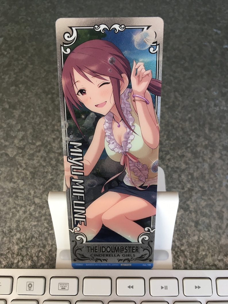 May include: A silver card featuring a cartoon image of a woman with long brown hair wearing a green and white dress. The text "THE IDOLM@STER CINDERELLA GIRLS" is on the card. The card is on a white keyboard.