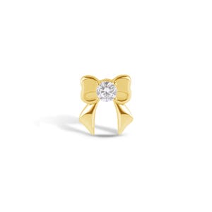 May include: A gold-toned bow-shaped jewelry piece featuring a single round, clear, faceted gemstone set in the center. The bow has smooth, polished surfaces with two downward-pointing tails and two rounded loops, creating a symmetrical design.