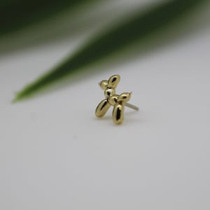 Puede incluir: Un pendiente de bot&oacute;n de perro globo de color dorado. El pendiente es una peque&ntilde;a y detallada escultura de un perro, con un poste para perforar. El pendiente est&aacute; sobre un fondo blanco con una hoja verde borrosa en el fondo.