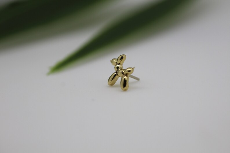 Puede incluir: Un pendiente de bot&oacute;n de perro globo de color dorado. El pendiente es una peque&ntilde;a y detallada escultura de un perro, con un poste para perforar. El pendiente est&aacute; sobre un fondo blanco con una hoja verde borrosa en el fondo.