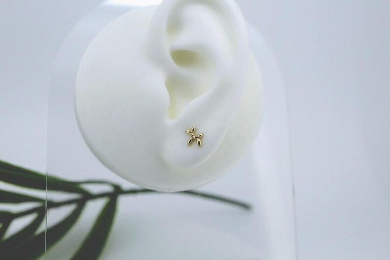 Puede incluir: Un pendiente de bot&oacute;n con forma de perro globo de color dorado se muestra en un modelo de oreja blanca. El pendiente es peque&ntilde;o y detallado, mostrando la forma ic&oacute;nica de un animal globo. El fondo es blanco suave y neutro.