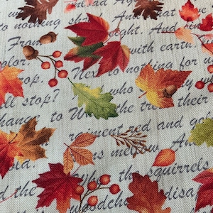 May include: Fabric with a fall theme, featuring colorful leaves in shades of red, orange, and yellow, along with acorns and berries. The fabric has a light background with script text.