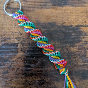 May include: A colorful keychain with a braided design. The keychain features a silver ring and a twisted braid of rainbow-colored rubber bands. The braid ends in a knot with several strands of colorful rubber bands. The keychain is on a dark wood surface.