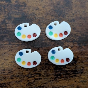 May include: Four white, paint palette-shaped charms with a hole at the top. Each charm has a series of colorful dots representing paint colors: blue, green, yellow, orange, and red. The charms are on a dark brown wooden surface.