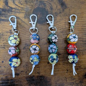 May include: Four keychains with decorative, painted ceramic beads. Each keychain has three colorful beads with floral designs, attached to a silver-colored clasp. The beads are in shades of yellow, pink, blue, and red.