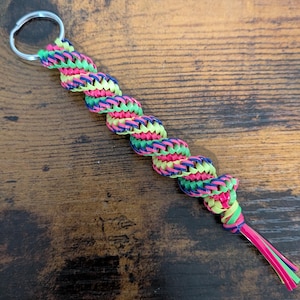 May include: A spiral keychain crafted from braided plastic cord in neon pink, lime green, and dark blue. The twisted, multi-colored lanyard is attached to a circular silver-toned metal split ring and ends with short, loose cord strands.