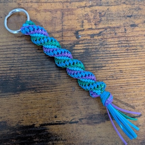May include: A keychain featuring a spiral-braided plastic cord in shades of purple, teal, and blue. The braided section is attached to a silver-colored metal split ring at one end and finishes with loose, dangling plastic strands at the other.