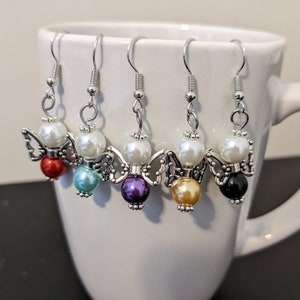 May include: Five pairs of angel-shaped earrings with silver-tone metal wings and a white pearl bead. Each pair features a different colored bead: red, teal, purple, gold, and black.