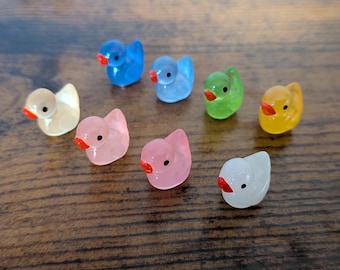 May include: A collection of small, colorful duck figurines. The ducks are in various colors, including blue, green, yellow, pink, and white, each with a red beak. These miniature ducks are likely decorative items or charms.