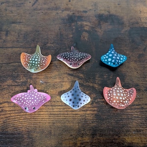 May include: Six colorful stingray figurines. Each figurine has a unique color scheme, including orange, purple, blue, and pink, with white polka dots. The figurines are arranged on a dark wooden surface.