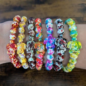 May include: Seven colorful bracelets with a turtle charm on each. The bracelets are made of white beads with colorful paint splatters. The turtles are painted in different colors, including red, yellow, brown, orange, pink, purple, and green.