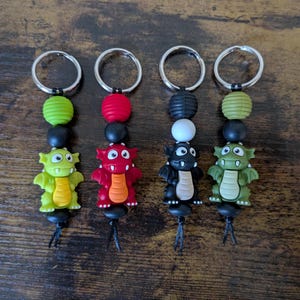 May include: Four colorful dragon keychains with different colored beads and a silver key ring. The dragons are green, red, black, and green.