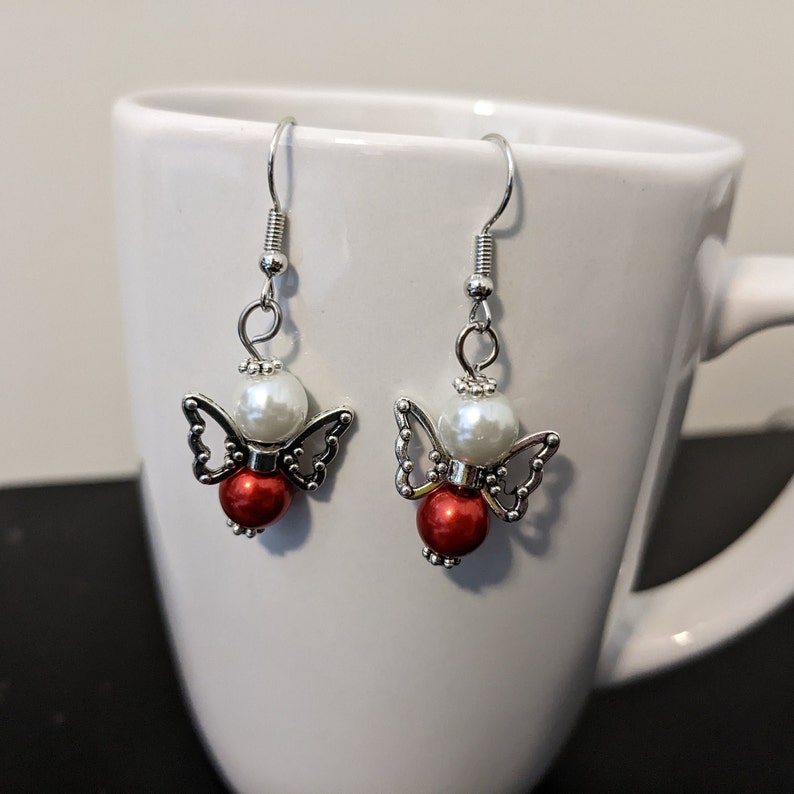 May include: A pair of silver angel-shaped earrings with white and red beads. The earrings are hanging from a white mug.