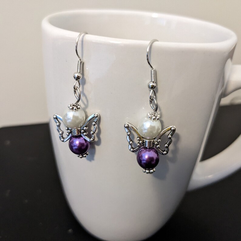 May include: A pair of silver angel-shaped earrings with white and purple beads.