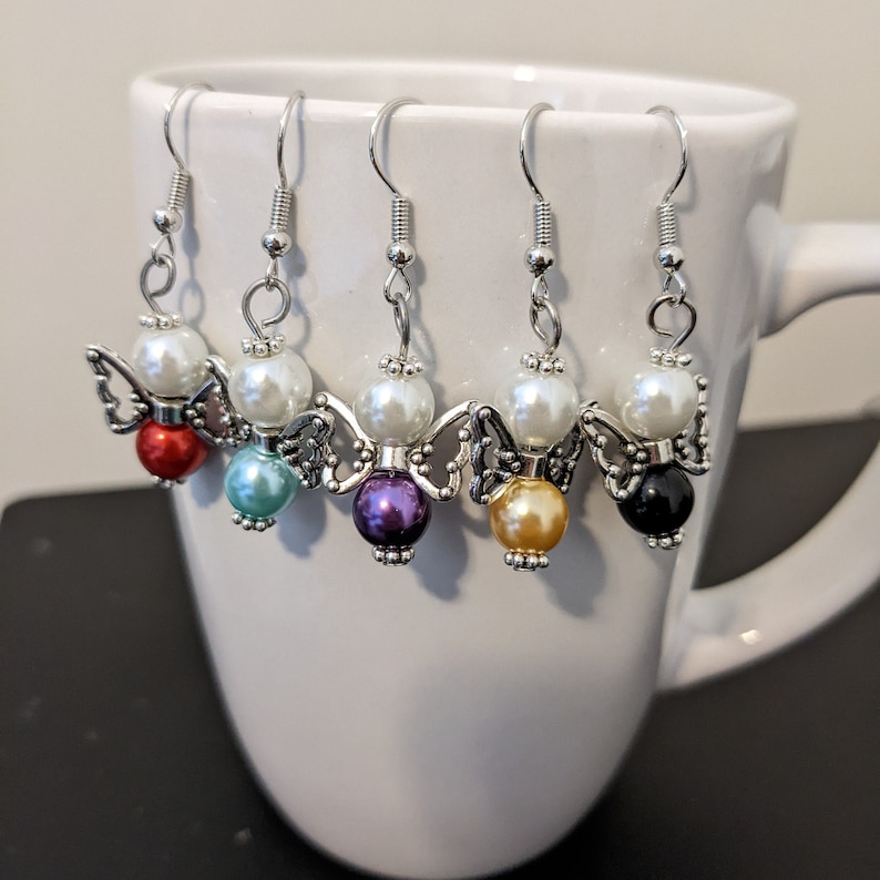May include: Five pairs of angel-shaped earrings with silver-tone metal wings and a white pearl bead. Each pair features a different colored bead: red, teal, purple, gold, and black.