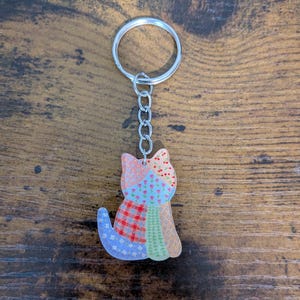 May include: A colorful cat-shaped keychain with a patchwork design. The keychain features a silver metal ring and chain. The cat's body is made up of various fabric patterns in shades of blue, red, green, and orange.