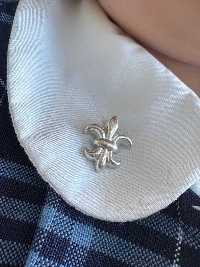 Puede incluir: Un broche de plata con forma de flor de lis sobre un cuello blanco.