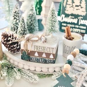 以下が含まれることがあります： ミニチュアのクリスマスツリー、松ぼっくり、シナモンスティック、そして「フレッシュカットクリスマスツリー」と書かれた木製看板で飾られた白い3段トレーです。