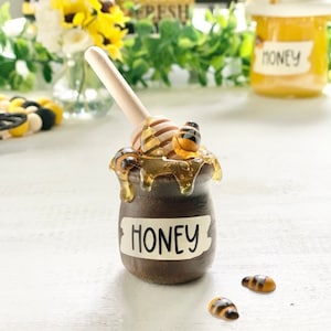 Może przedstawiać: Mały brązowy słoik miodu z drewnianym nabierakiem do miodu i pszczołami na górze. Słoik ma białą etykietę z napisem "Miód".