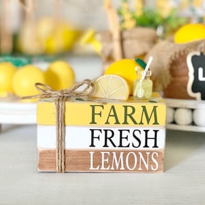 以下が含まれることがあります： 「FARM FRESH LEMONS」と描かれた3つの木製のブロックの積み重ね。ブロックは、レモンのスライス、レモン全体、ストローが付いた小さなガラス瓶で飾られています。