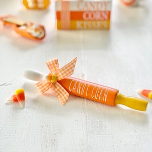 Può includere: Un mattarello arancione con le parole "CANDY CORN" in lettere bianche, legato con un fiocco a quadretti. Il mattarello ha manici gialli ed è circondato da pezzi di caramelle a forma di mais su una superficie bianca. Sullo sfondo c'è una scatola con le parole "CANDY CORN KISSES".