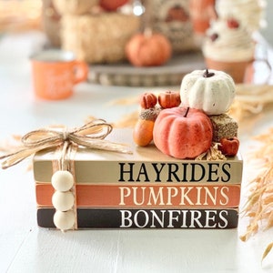 May include: A stack of three decorative wooden books tied with twine and adorned with wooden beads. The books are painted in neutral tones and feature the words "HAYRIDES", "PUMPKINS", and "BONFIRES". Miniature pumpkins and acorns sit atop the books.